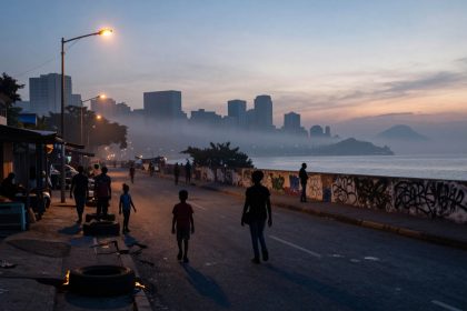 People walk along a waterfront street at dusk, with silhouetted skyscrapers in the background. Streetlights illuminate the scene, and graffiti covers a low wall by the water. Smoke or fog partially obscures the distant buildings.