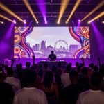 A DJ performs on stage with a vibrant digital backdrop featuring abstract patterns and a cityscape, including a Ferris wheel. The crowd in the foreground is silhouetted against purple and yellow stage lights.