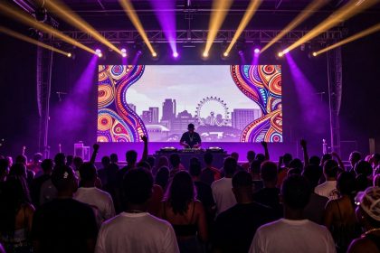 A DJ performs on stage with a vibrant digital backdrop featuring abstract patterns and a cityscape, including a Ferris wheel. The crowd in the foreground is silhouetted against purple and yellow stage lights.