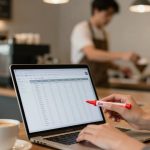 A person holds a red pen, pointing at a laptop displaying a spreadsheet in a cafe. A cup of coffee sits nearby. A barista works in the background.