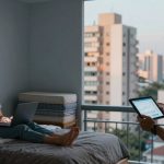 A woman sits on a bed using a laptop, while a man stands by a window holding a tablet displaying graphs. The room has a gray wall and large windows showing city buildings.
