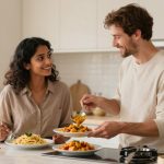A woman and a man are in a kitchen. The woman is holding a fork by a plate of spaghetti, smiling at the man. The man is serving pasta with vegetables onto plates. Various jars are on the counter, and the kitchen is bright and modern.