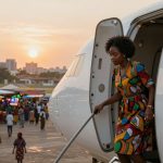 A woman in a colorful patterned dress descends the stairs of an airplane. In the background, a market with stalls and people is visible at sunset.