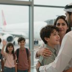 A family at an airport, with a man in traditional attire hugging a crying boy. A woman stands beside them, and three children wave in the background. An airplane is visible through the large windows.