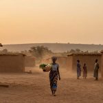 A woman walks through a dusty village pathway carrying a basket of vegetables on her head. She wears a patterned skirt and headscarf. Mud brick houses and three people are in the background, with a hazy sunset sky and distant hills.