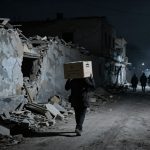 A person carrying a cardboard box on their head walks through a dimly lit, debris-filled street with damaged buildings. Shadows and silhouettes of other people are visible in the background.