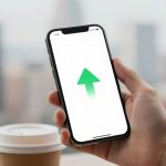 A hand holds a smartphone displaying a green upward arrow on a white screen. A coffee cup with a lid is in the blurred background.