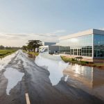 A flooded road with three people standing on the left. A modern building with glass windows and solar panels is on the right. Fields and trees are in the background. A translucent world map overlay covers the scene.