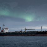 A large cargo ship with a rusted hull sails on a dark ocean. The sky above has a faint green aurora, and icy mountains are visible in the background.