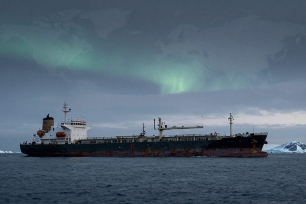 A large cargo ship with a rusted hull sails on a dark ocean. The sky above has a faint green aurora, and icy mountains are visible in the background.