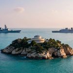 A small island with a circular building and greenery is in the foreground. Two naval ships sail parallel in the background on a calm sea.
