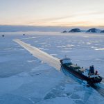 A large cargo ship navigates through icy waters, creating a path in the frozen sea. It is surrounded by ice floes, with distant snow-covered mountains under a pastel sky.