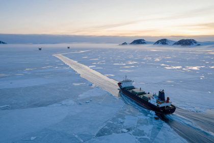 A large cargo ship navigates through icy waters, creating a path in the frozen sea. It is surrounded by ice floes, with distant snow-covered mountains under a pastel sky.