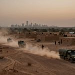 Convoy of military trucks driving on a dusty desert road through a village with sand-colored buildings at dusk. City skyline visible in the distance.