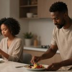 A man and woman in a kitchen. The man is preparing a salad on a plate, while the woman, seated, looks at her phone. Both wear neutral-toned clothing.