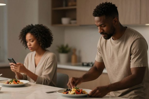 A man and woman in a kitchen. The man is preparing a salad on a plate, while the woman, seated, looks at her phone. Both wear neutral-toned clothing.