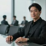 A person in a black suit sits at a conference table, smiling, with blurred colleagues in business attire in the background.
