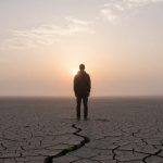 A person stands on cracked, dry earth facing a hazy sunset. The ground is barren with a visible crack leading towards them.
