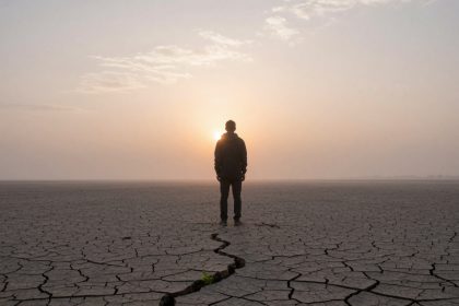 A person stands on cracked, dry earth facing a hazy sunset. The ground is barren with a visible crack leading towards them.