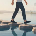 A person in black shoes and rolled-up jeans walks across stepping stones on water. A large calendar is visible in the background.