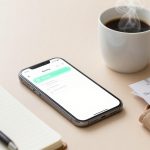 A smartphone displaying an earnings app, a steaming cup of coffee, a pen on an open notebook, and a wallet with receipts on a beige surface.