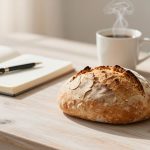 A round loaf of crusty bread on a wooden table with an open notebook and pen. A steaming cup of coffee and a small potted succulent are in the background.