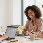 Three people are sitting at a table with laptops, collaborating. A woman on the left types, while a woman in the middle and a man on the right take notes. A cup and sticky notes are on the table.