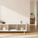 White shelving unit with books, a vase, and a mug on top. A woven basket and coiled cable are also placed on it. A hallway with a table and vase of flowers is visible in the background. Light wood flooring and a white rug are in the foreground.