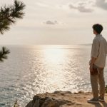 A person stands on a rocky cliff holding a book, gazing at the sunlit ocean. Pine trees frame the scene under a partly cloudy sky.