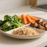 A divided plate on a kitchen counter holds leafy greens, carrot sticks, sesame-coated snacks, and puffed grains. Walnuts are nearby.