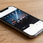 A smartphone displaying a video editing app on its screen, showing a musician performing on stage under bright lights, placed on a wooden surface.