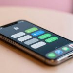 A smartphone lies on a light wooden table displaying a screen with various messaging app icons and notifications in green and blue.