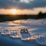 A gold ring rests on a white lace cloth with intricate patterns. The background features a sunset with a soft, cloudy sky.