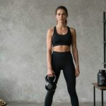 A woman in black athletic wear holds a kettlebell in a minimalist gym. Shelves with supplements are on the wall, and a rope is coiled on the floor.