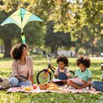 A family sits on a red checkered picnic blanket in a park. Two adults and two children are engaged in conversation and drawing. A bicycle and kite are nearby, with trees in the background.