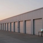 A row of storage unit doors on a beige metal building at sunset. A lighthouse is visible in the background. A small table with a glowing tablet stands on the concrete walkway.