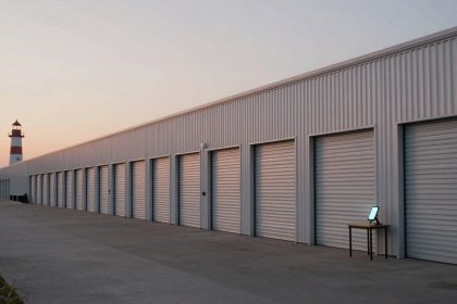 A row of storage unit doors on a beige metal building at sunset. A lighthouse is visible in the background. A small table with a glowing tablet stands on the concrete walkway.