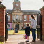 A woman and a child stand in front of an open wrought iron gate leading to a brick school building with a clock tower. The child, wearing a school uniform and holding a colorful lunchbox, looks towards the school. The woman, in casual attire, stands beside the child. A chalkboard sign is visible on the path.
