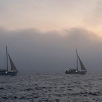 Two sailboats with lit lanterns on a calm sea at dusk, under a cloudy sky with a subtle glow from the setting sun.