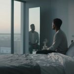 A person sits on a bed in a softly lit room, holding a steaming cup. A large window reveals a cityscape at dawn. A mirror reflects the scene.