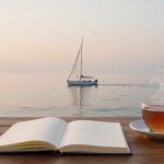 An open book and steaming cup of tea on a wooden table with a sailboat on calm water at sunset in the background.