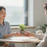 A woman in a blue shirt hands a small potted plant with steam rising from it to a man in a white shirt. They sit at a round table in a bright room with a window and a teapot in the background.
