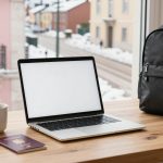 A laptop with a blank screen sits on a wooden table next to a steaming mug, a passport, and a black backpack. Snowy buildings are visible through the window.