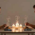 A couple sits across from each other at a table, enjoying a candlelit dinner. Steam rises from their food, with smartphones placed beside their plates.