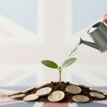 A hand waters a small green plant with a silver watering can. The plant is growing from a pile of soil and British coins, with a blurred Union Jack flag in the background.
