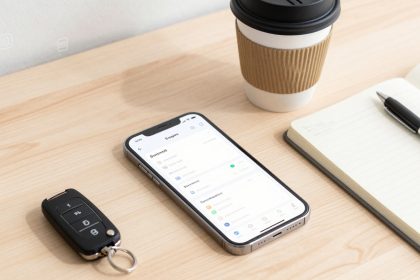 Smartphone displaying an app lies on a wooden desk next to a car key, a lined notebook with a pen, and a takeaway coffee cup.
