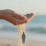 A hand releasing a stream of sand onto a beach, with the ocean in the background. A silhouette of a person is superimposed on the sand.
