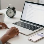 A person works at a laptop on a white desk, with a white coffee mug, smartphone, and two black alarm clocks nearby.