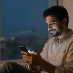 A man sitting indoors at night, smiling at a smartphone. He is wearing a light-colored shirt. A window shows a blurred cityscape.