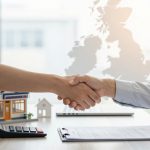 Two people in business attire shake hands over a desk with a contract, calculator, and model houses. A map is visible in the background.